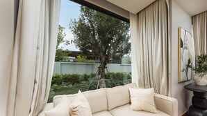 Ein modernes Wohnzimmer mit einem beigefarbenen Sofa, einem großen Fenster mit Vorhängen und Blick auf einen Baum und Grünfläche.