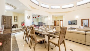 Une salle à manger spacieuse avec un plafond haut et de nombreuses fenêtres, une cuisine moderne en arrière-plan et un salon confortable avec un canapé et une télévision.