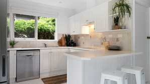 Una cocina moderna con muebles blancos, un lavavajillas Bosch, un ventanal con vegetación a la vista y una encimera con utensilios de cocina.