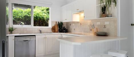 Una cocina moderna con muebles blancos, un lavavajillas Bosch, un ventanal con vegetación a la vista y una encimera con utensilios de cocina.