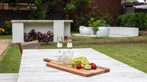 Mesa de comedor al aire libre con una tabla de madera que sostiene una botella, dos vasos y un racimo de uvas.