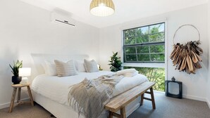 Un dormitorio con una cama grande, una mesita de noche con una lámpara, una ventana con vistas a árboles y una planta en la pared.