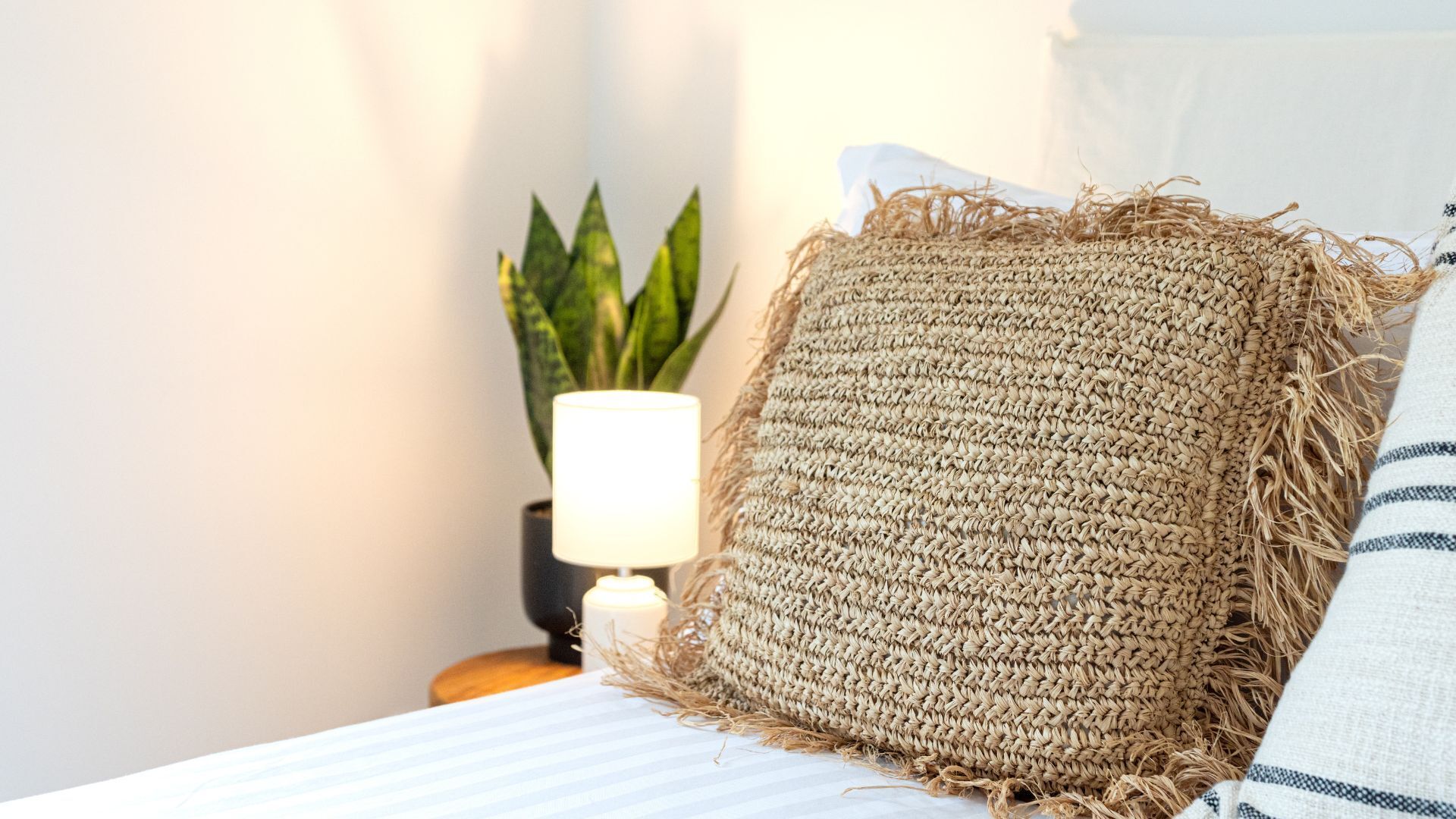 A bed with a striped pillow and a woven throw. There is a lamp on a bedside table and a plant in the background.