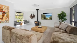 A modern living room with a fireplace, comfortable seating, a large window, and a painting on the wall. - Kabina 98 – coastal beach cottage (Callala Beach)