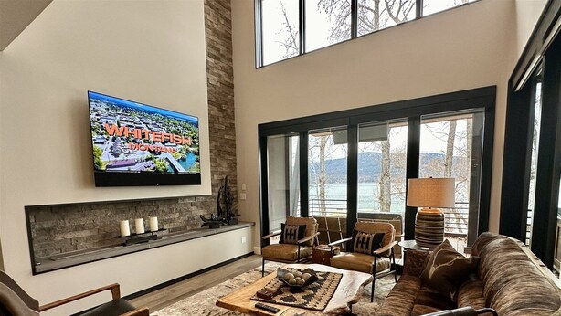 Uma sala de estar ampla com uma televisão grande fixada na parede, lareira e vista para um lago através de grandes janelas.