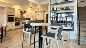 Cozinha moderna com eletrodomésticos embutidos, sala de jantar com mesa redonda e cadeiras, e estante com objetos decorativos.