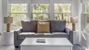 Un salon avec un canapé gris, une table basse blanche et un tapis à motifs. Il y a deux fenêtres avec des rideaux, un ventilateur de plafond et des lampes décoratives.