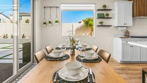 A dining area with a wooden table set for six, surrounded by chairs. There is a window with a view of the outside, a kitchen area with cabinets and a countertop, and a wall-mounted plant holder with three plants. - New Pool+Spa+Games+Large BBQ+Designer+Beautiful (Coachella)