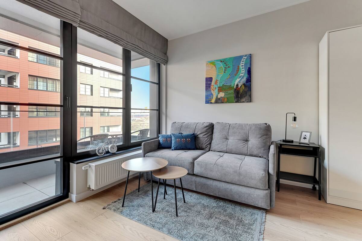 A modern living room with a grey sofa, a small round table, and a large window with a view of buildings.