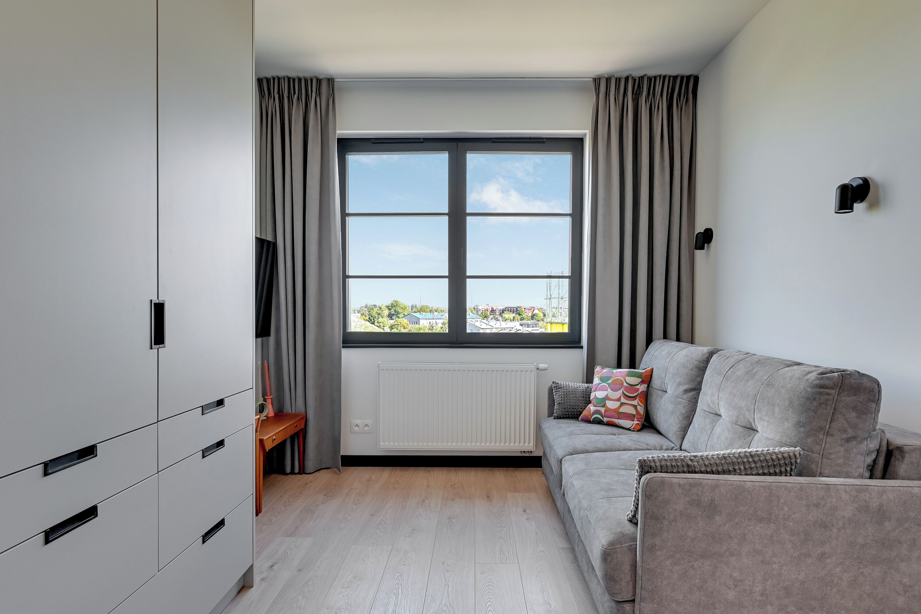 Un salon moderne avec un canapé gris, une table d’appoint en bois et une grande fenêtre avec des rideaux.