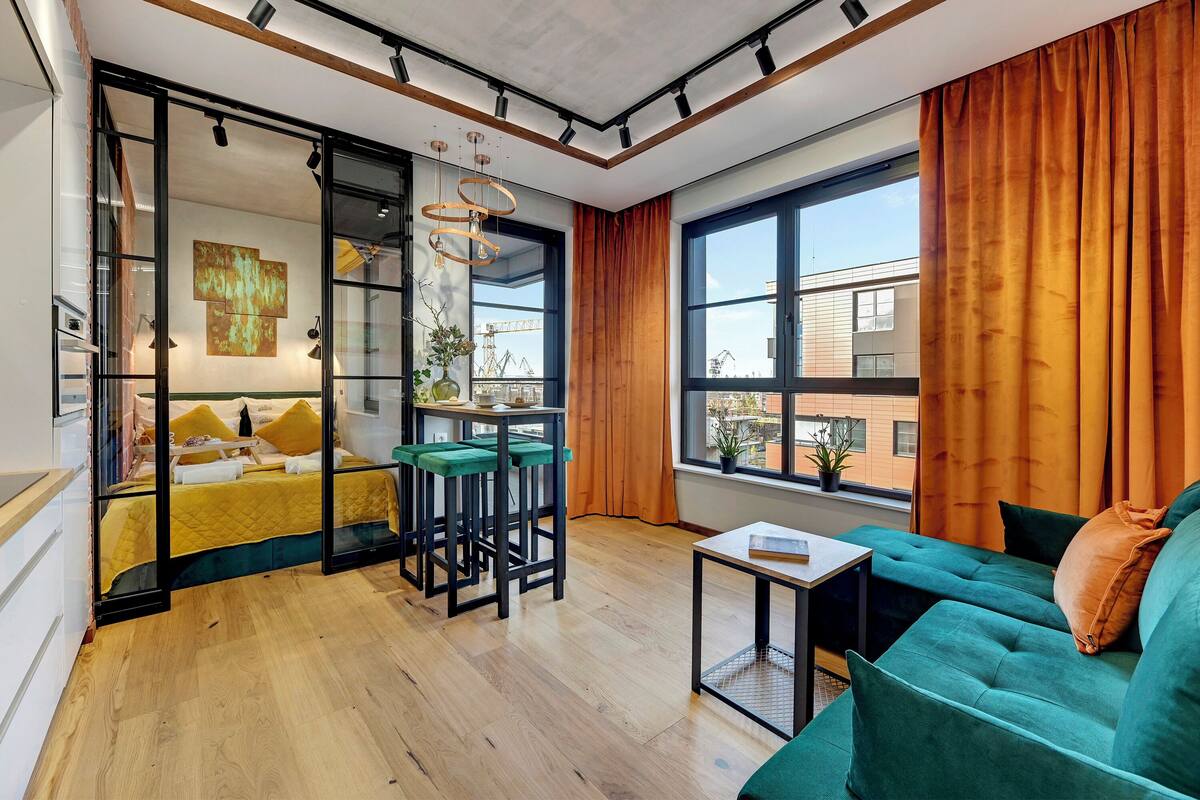 A modern living room with a sofa, a small table, a bed with a yellow bedspread, and a glass partition with a view of the outside.