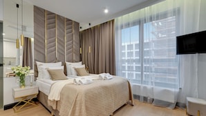 A modern bedroom with a large bed, a bedside table, a TV, and a view of the outside. - Doki Living & Foodhall by Downtown Apartments (Gdansk)