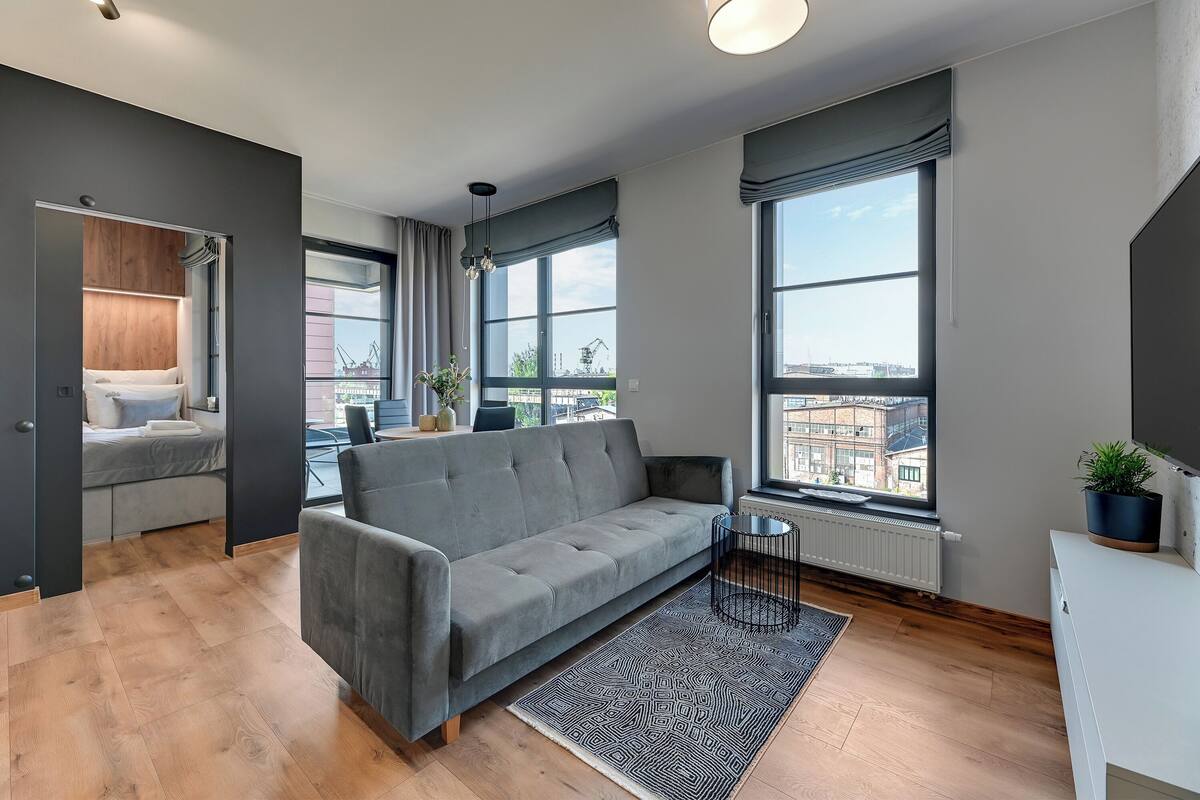 A modern living room with a grey sofa, wooden flooring, large windows, and a view of buildings.
