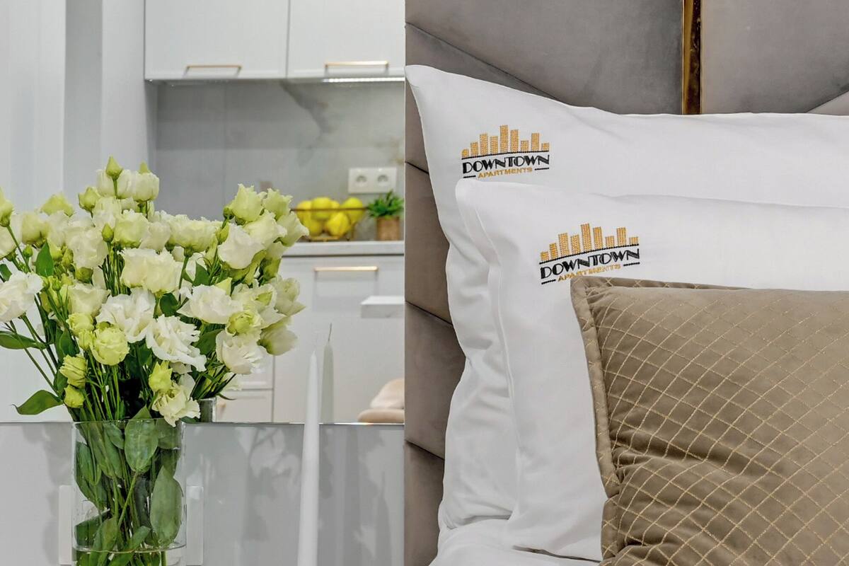 A bedroom with a bed, a nightstand, a vase of flowers, and a view of the kitchen.