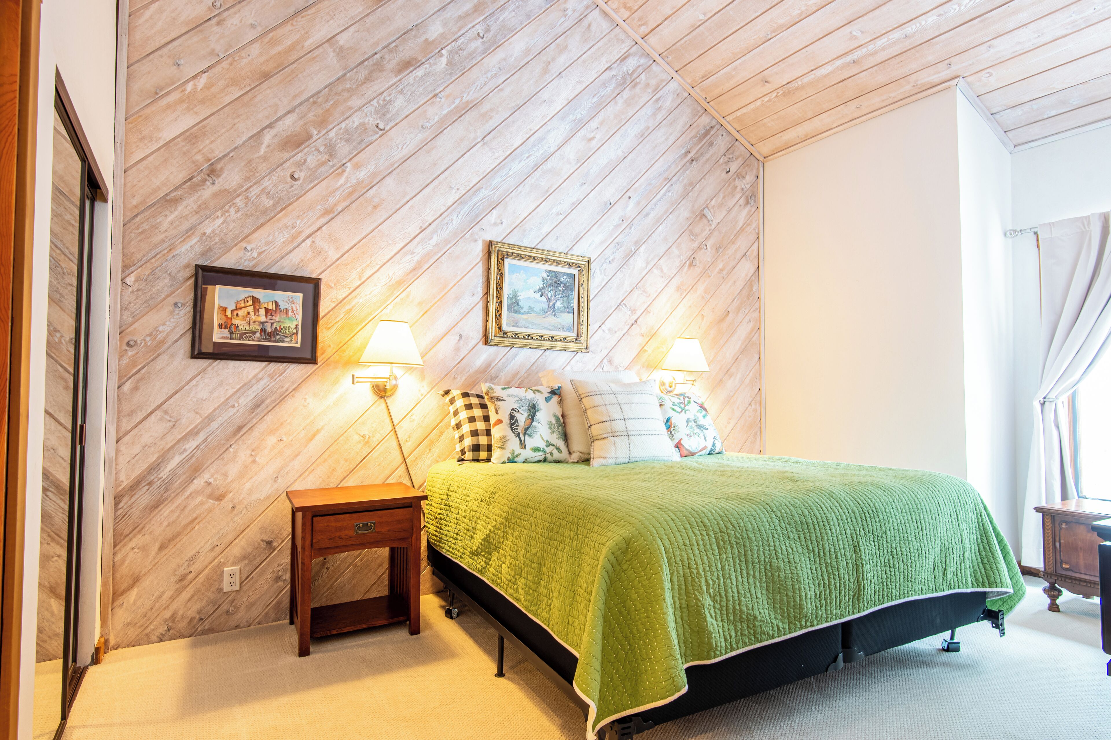 A bedroom with a wooden sloped ceiling, a green bedspread, two bedside lamps, and two framed pictures on the wall.