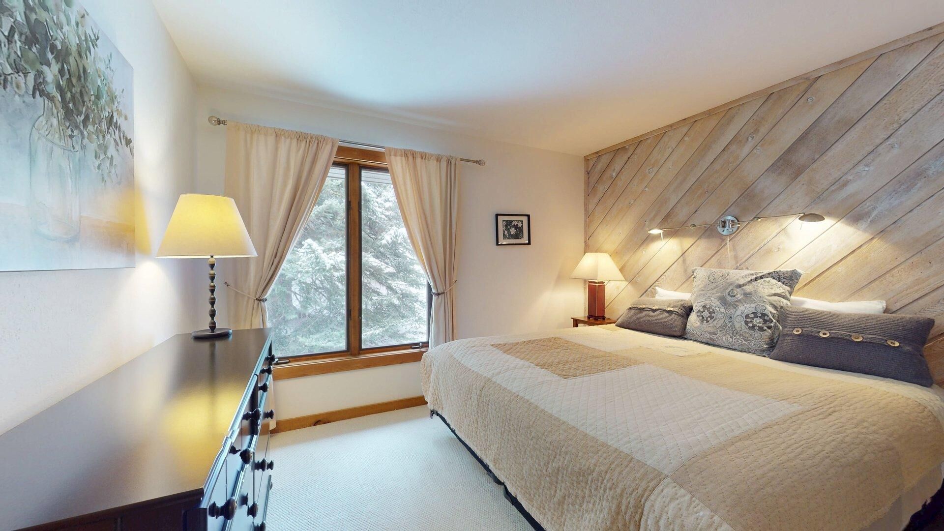 A bedroom with a bed, two bedside lamps, a window with curtains, a framed picture on the wall, and a guitar leaning against the wall.