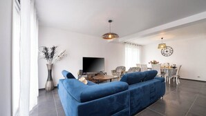 A modern living room with a blue sofa, a flat-screen TV, a dining table with chairs, and a decorative vase.