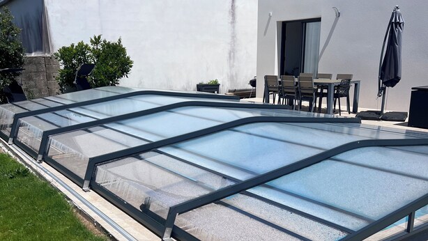 A swimming pool with a glass enclosure, a patio area with outdoor furniture, and a white building with a stone foundation.