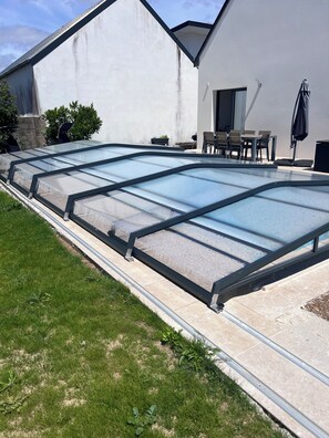 A swimming pool with a glass enclosure, a patio area with outdoor furniture, and a white building with a stone foundation.