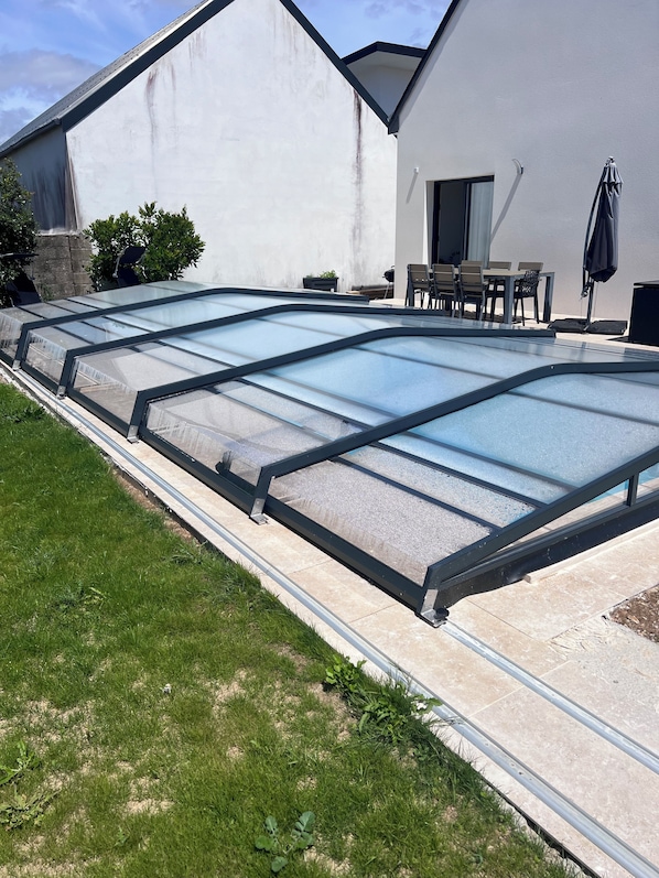 A swimming pool with a glass enclosure, a patio area with outdoor furniture, and a white building with a stone foundation.