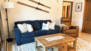 Ein Wohnzimmer mit einem blauen Sofa, einem Holztisch, einem braunen Ledersessel und einem Teppich mit Muster.