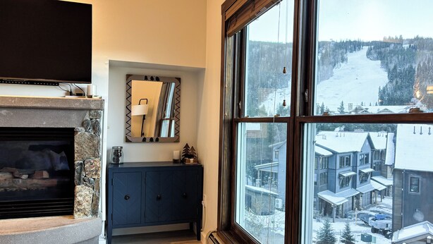 Ein Wohnzimmer mit Kamin aus Stein, einem dunkelblauen Schrank und einem großen Fenster mit Blick auf ein verschneites Dorf.