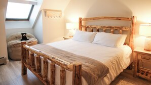 Ein Schlafzimmer mit schrägverdachter Decke, einem Bettgestell aus Holz, weißer Bettwäsche, einer Nachttischlampe und einem kleinen Stofftier auf einem Sitzsack.