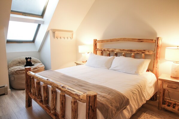 A bedroom with a sloped ceiling, a wooden bed frame, white bedding, a bedside lamp, and a small stuffed animal on a bean bag chair.