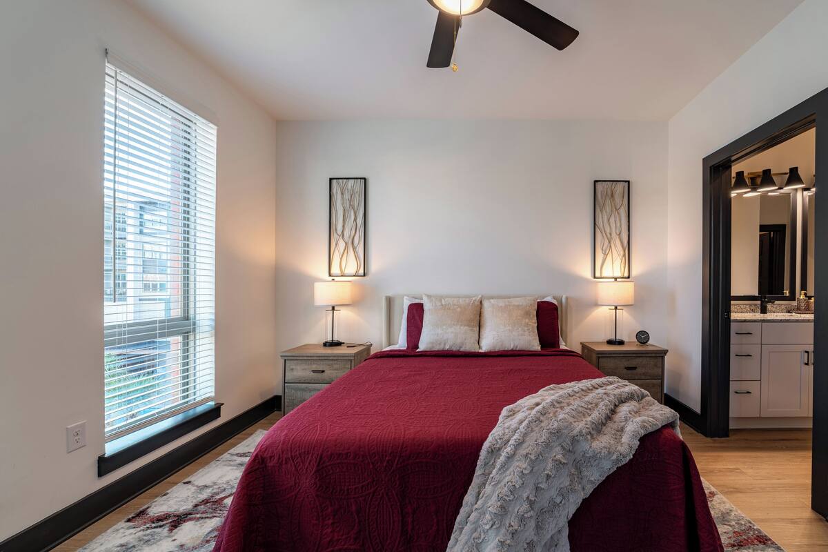 A bedroom with a large bed, two bedside tables with lamps, a ceiling fan, a window with blinds, and a bathroom visible through an open door.