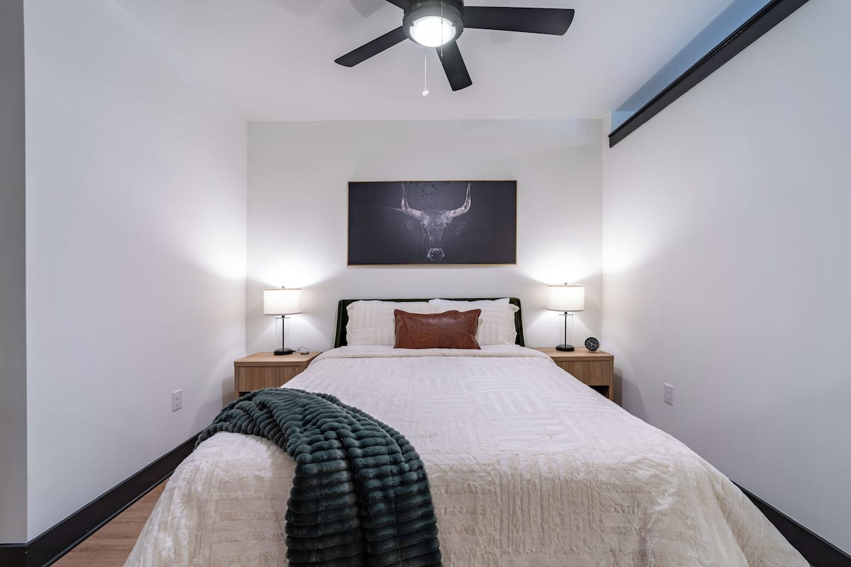 A bedroom with a bed, two bedside lamps, a ceiling fan, and a framed artwork on the wall.