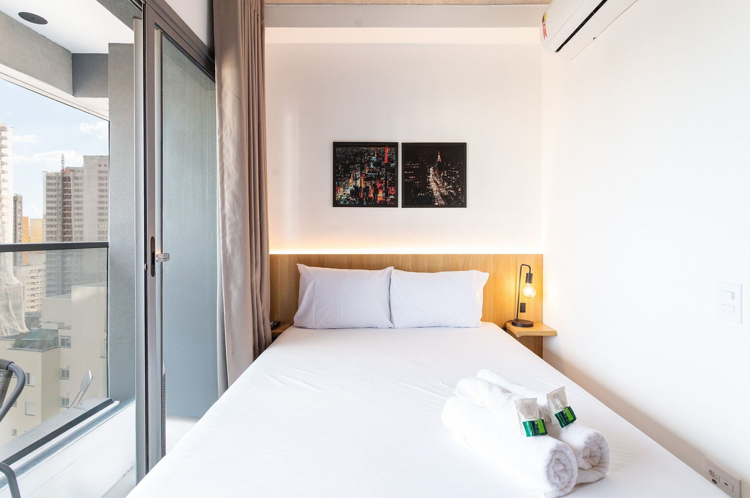 Ein Hotelzimmer mit einem Bett, zwei gerahmten Bildern an der Wand, einem Nachttisch mit einer Lampe und einem Fenster mit Blick auf Gebäude.