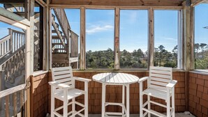 Un balcón con dos sillas blancas y una mesa pequeña, barandillas de madera y vista a un área boscosa.