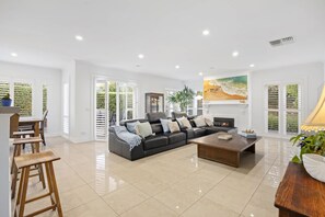 A spacious living room with a black sofa, a coffee table, and a fireplace. There are large windows with blinds, a dining area with a table and chairs, and a potted plant.