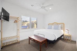 Une chambre à coucher comprenant un lit, une télévision, une fenêtre avec des stores, un ventilateur de plafond et une table de chevet.