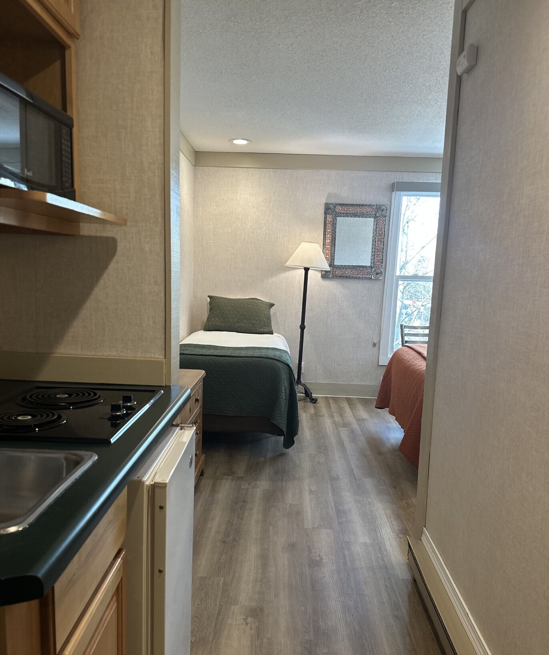 Une cuisine compacte équipée d’une cuisinière, d’un réfrigérateur et de meubles de rangement en bois. Un lit recouvert d’une couverture verte et agrémenté d’un oreiller est placé contre le mur, avec une lampe sur pied à proximité. Une fenêtre offre une vue sur les arbres.