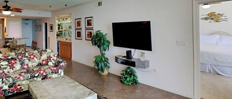 A living room with a sofa, coffee table, and two potted plants. There is a flat-screen TV mounted on the wall and a ceiling fan above.