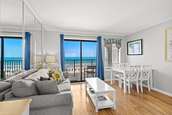 Una sala de estar moderna con sofá, mesa de centro y comedor, ventanas amplias con vista a la playa y un balcón con barandilla.