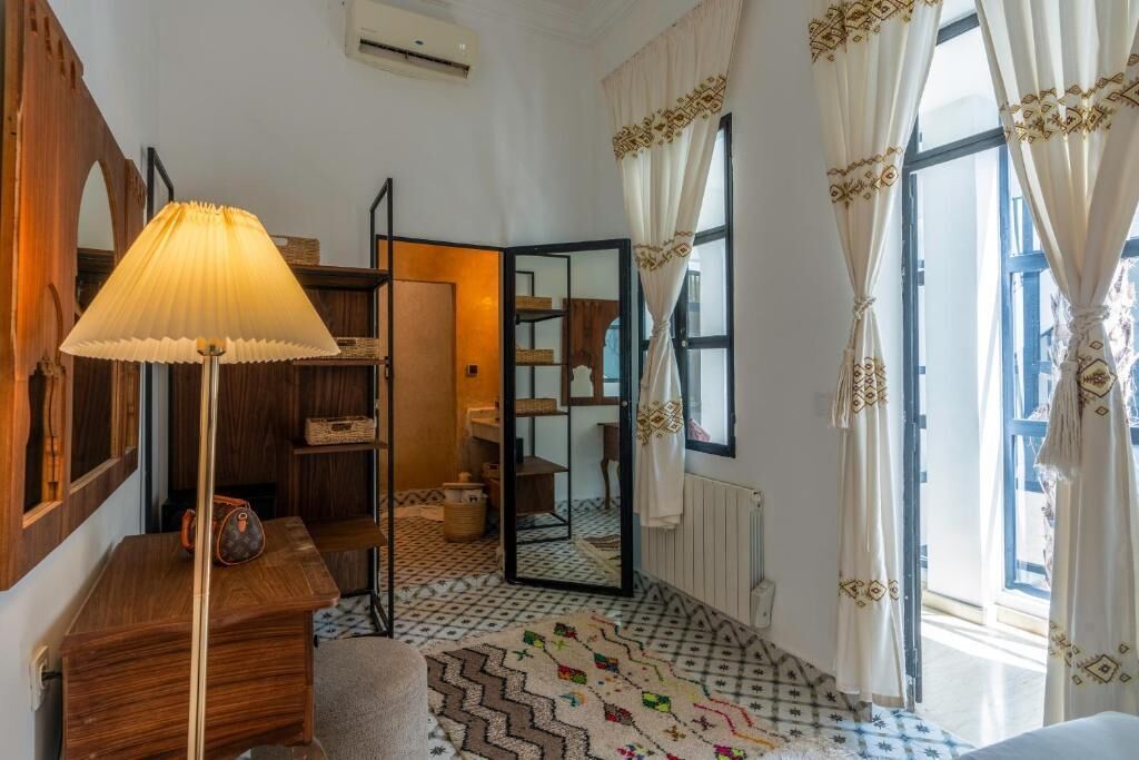 A hotel room with a wooden floor, a mirror, a lamp, a patterned rug, and a view of the outside through a window with curtains.