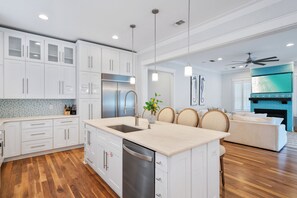 A modern kitchen with white cabinetry, a stainless steel refrigerator, and a dishwasher. There is a dining area with a white sofa and a ceiling fan. - Coconut Cove-Private Pool, Golf Cart, Bikes, Walk to Beach, Discount Rates. (Santa Rosa Beach)