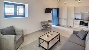 A modern living room with a sofa, a coffee table, a dining table with chairs, a kitchen area with cabinets and a TV, and a window with a view. - Charming sheepfold on the Liamone beach (Casaglione)