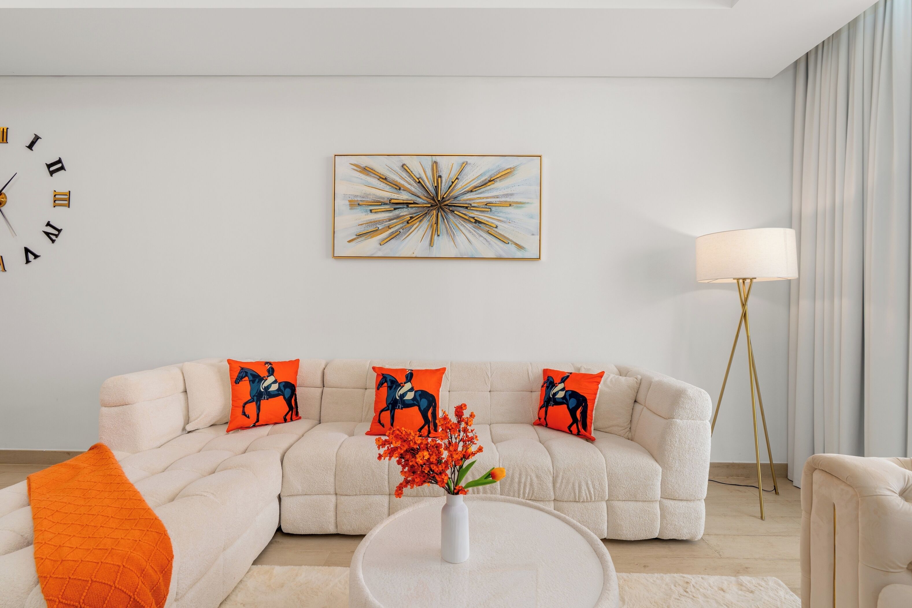A modern living room with a beige sofa, orange cushions, a round coffee table, a white vase with flowers, a floor lamp, and a wall clock.