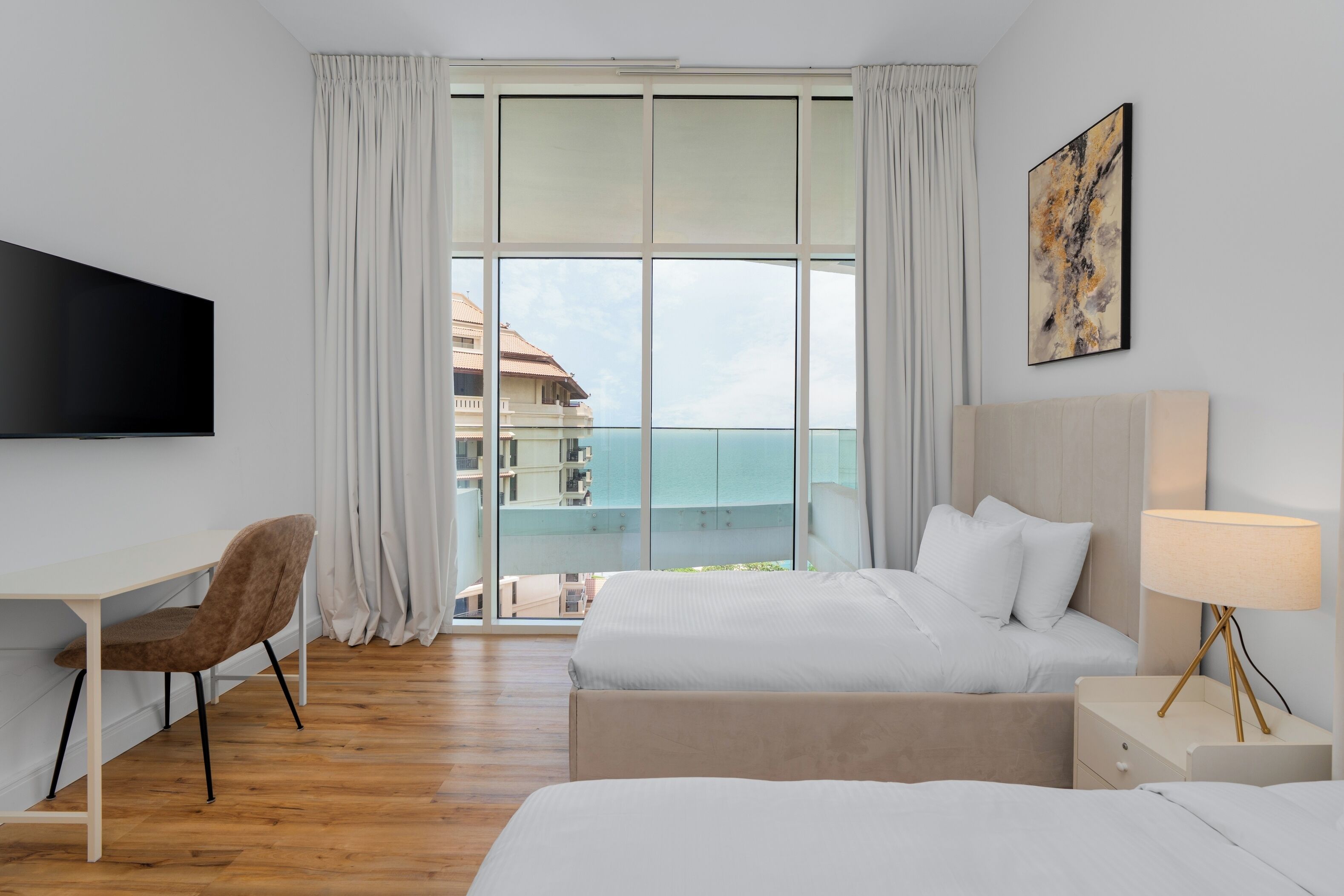 Habitación de hotel con cama, escritorio, silla, televisión, lámpara de noche y un amplio ventanal con vistas a edificios y al agua.