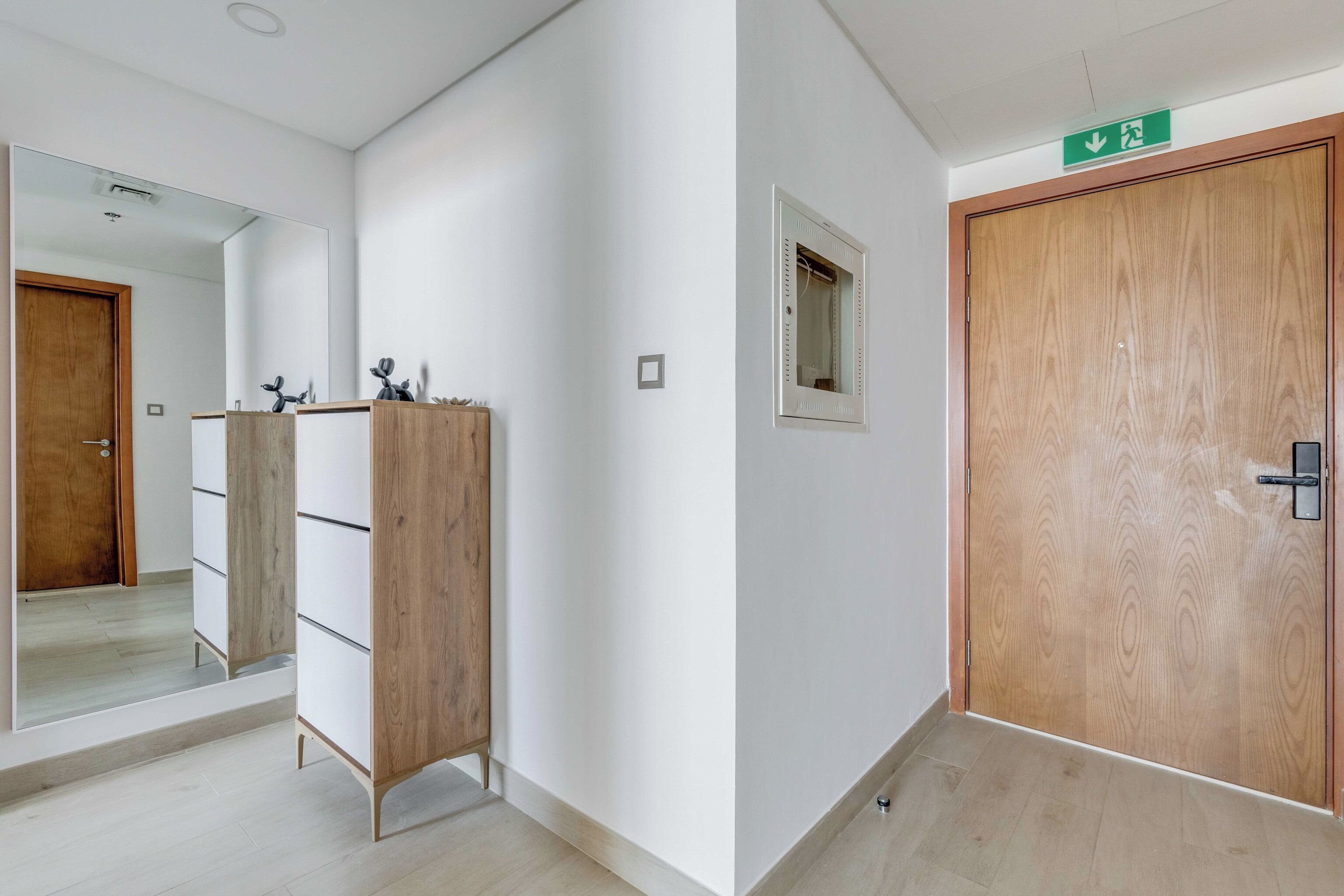 A hallway with a wooden wardrobe, a mirror, and a door with a green exit sign.