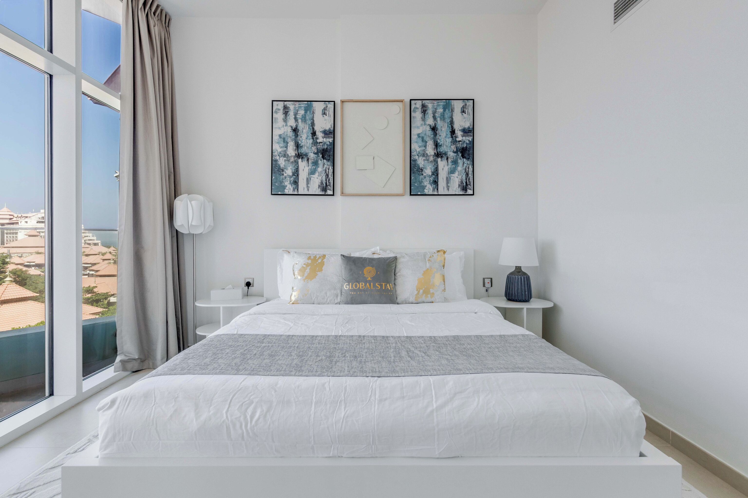 A modern hotel room with a large bed, a view of the city, and artwork on the wall.