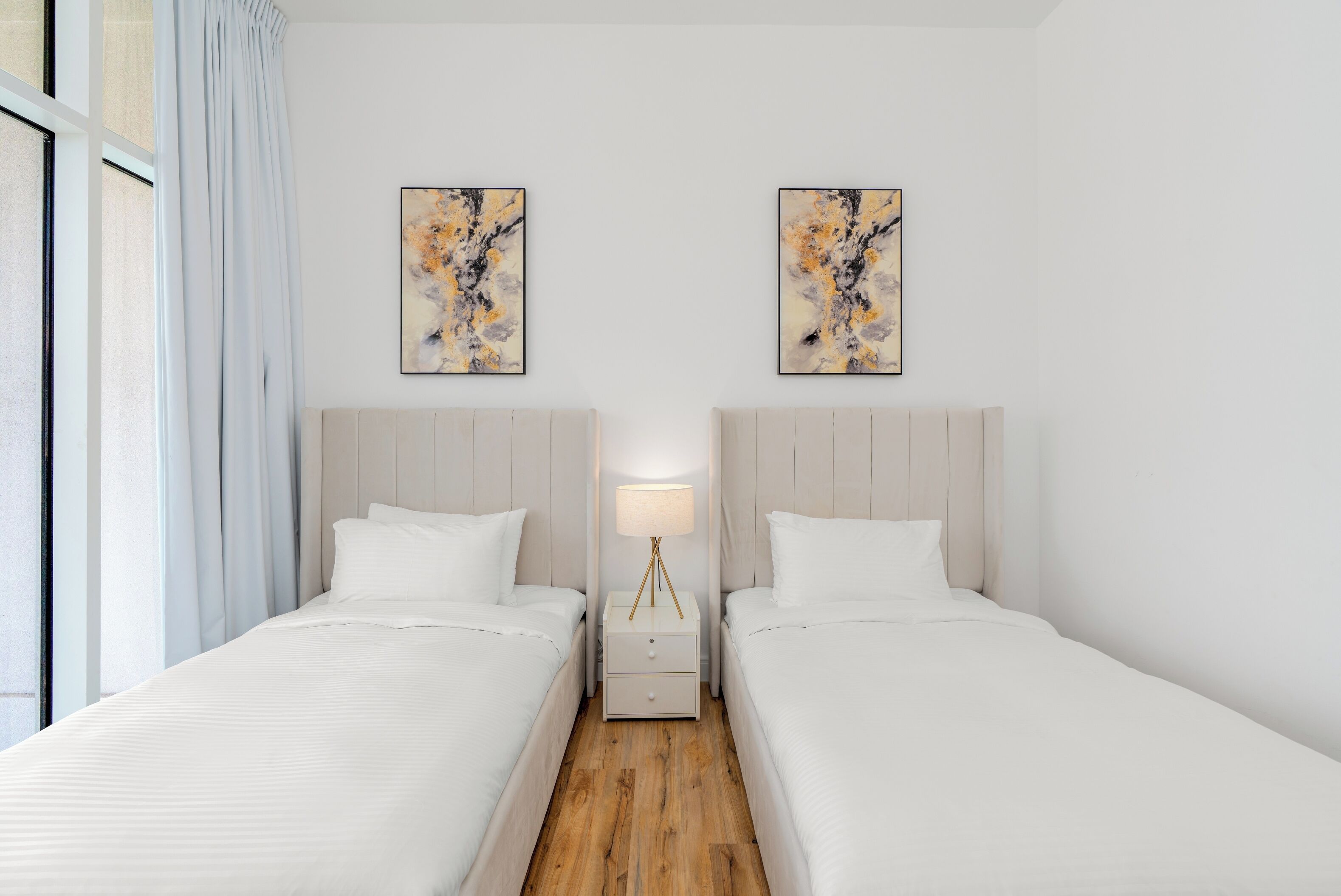 A hotel room with two beds, a nightstand with a lamp, and two abstract paintings on the wall.