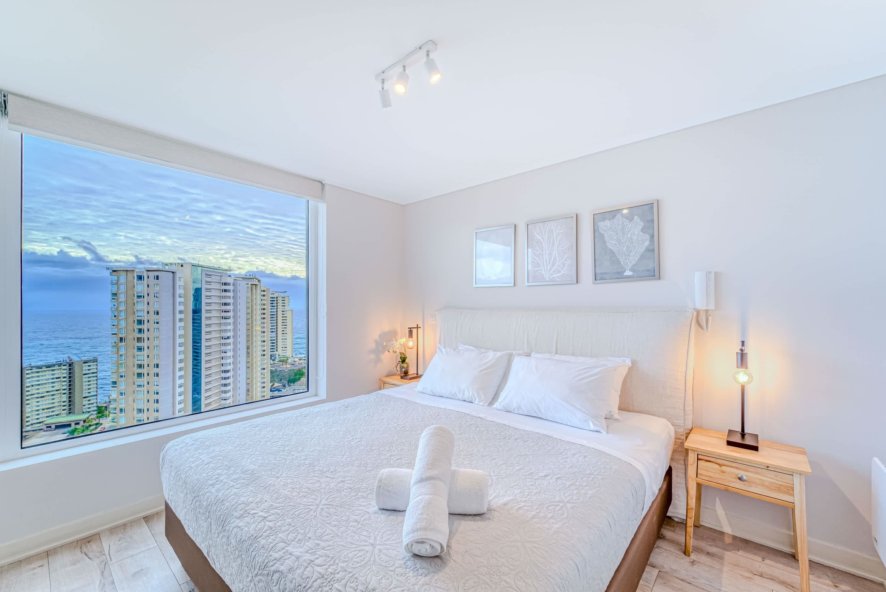 A hotel room with a large bed, a nightstand, a lamp, and a view of tall buildings through a large window.