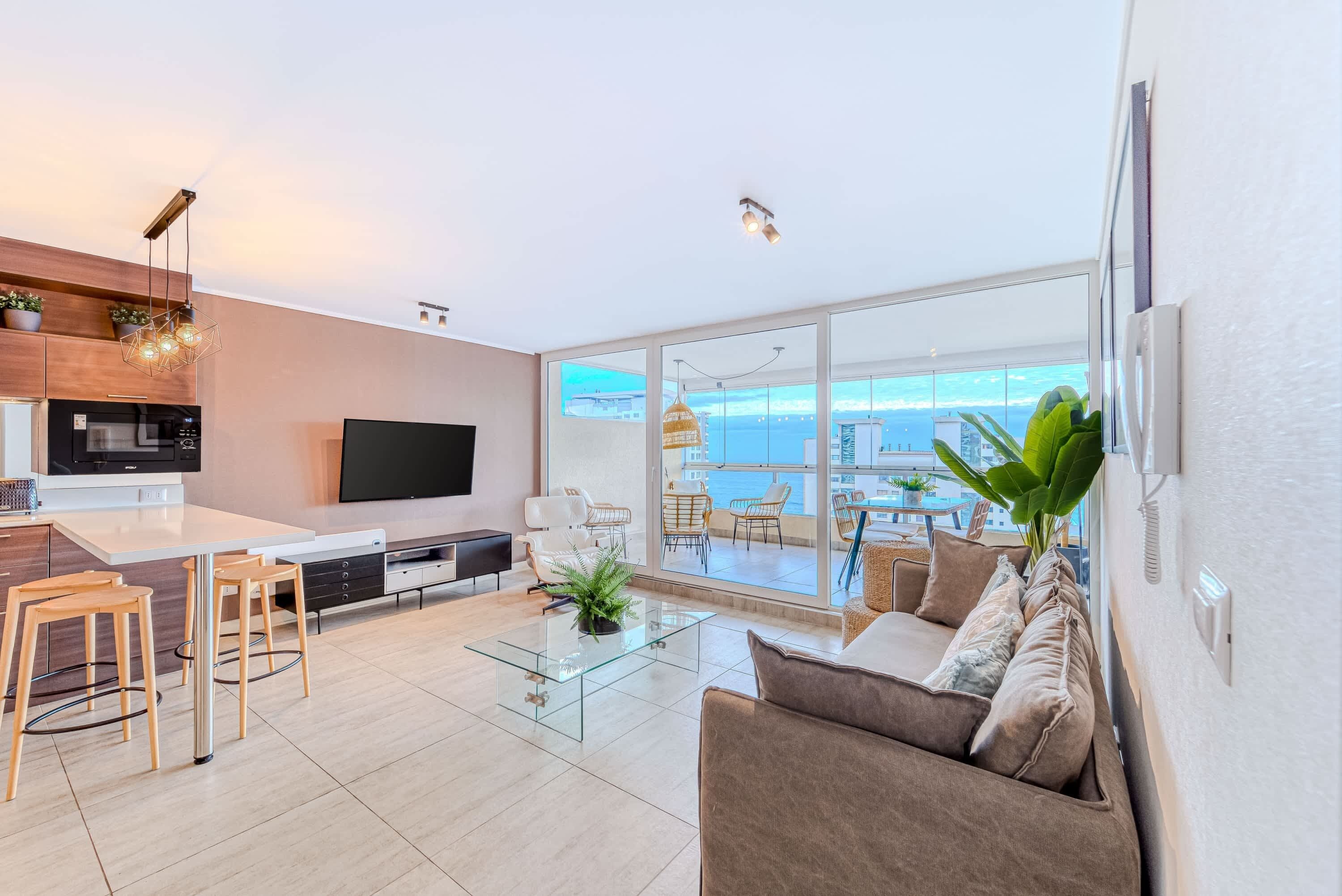 A modern living room with a large TV, a glass coffee table, and a sofa. There is a kitchen area with a microwave and a dining area with chairs.