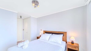 A neatly made bed with white linens and a wooden headboard, two bedside lamps, and a white door in the background. - Rincon Miramar (Concón)