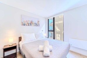 A hotel room with a bed, a nightstand with a lamp, a painting on the wall, and a window with a view of buildings.