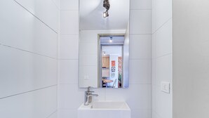 A modern bathroom with white tiles, a sink, a mirror, and a door leading to a room with a table and chairs. - Espacio Nunoa 1BR, balcony and free parking (Ñuñoa)
